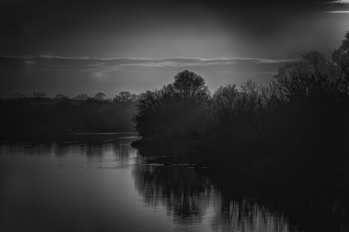 Czarno-białe nastrojowe zdjęcie ukazujące zakole rzeki z drzewami. 